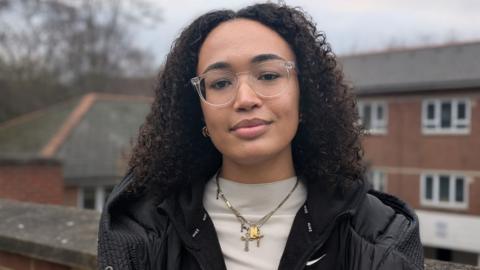 Olimata stands outdoors facing the camera and has long, curly black hair and glasses, wearing a black jacket, white top and jewellery around her neck