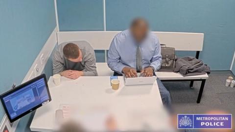 Man in grey sweater looks down to his right he is in a police custody room and man next t him, face blurred is typing on laptop
