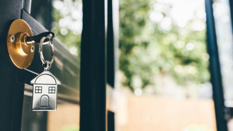A key in an opened door. The key has a house-shaped fob key ring. Bright light pours in through the open door.