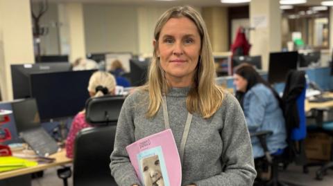 Sarah Terrett has long blonde hair and is looking into camera, smiling slightly. She's wearing a grey sweater and holding a pink booklet about pregnancy with a picture of a baby on. She's standing in an office, with colleagues sat behind at desks. The background is slightly blurred but she is in focus.