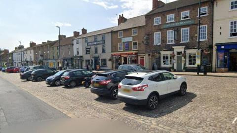 Bedale Market Place with parked cars, shops and pubs