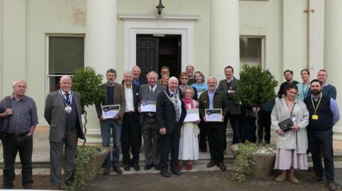 A group photograph showing the award winners and council dignitaries holding their certificates, standing outside a white building which has pillars.