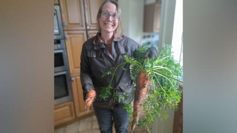 Diana McIntyre standing in her kitchen holding the giant carrot. She is smiling widely and is wearing a khaki waxed jacket and jeans. The carrot is covered in mud and has huge green fronds.