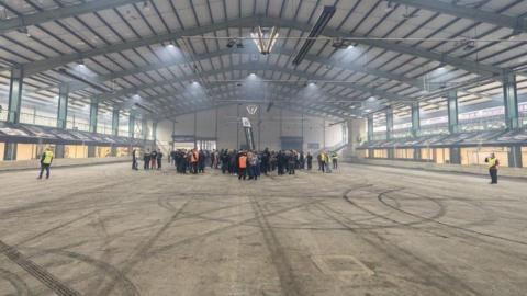 The inside of a large industrial unit, with people stood around in the centre. A series of tyre marks can be seen on the ground.