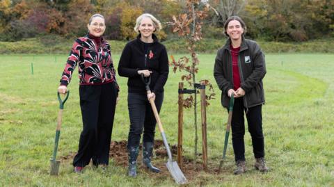 Three women are planting oak trees in a country park. They all have spades and are looking at the camera and smiling. There is a small oak tree planted next to them.