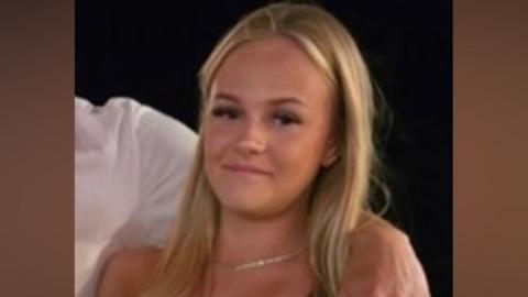 A slightly blurred image of a young girl, with long blonde hair, smiling at the camera. She is wearing a necklace and a person has their arm around her, who is wearing a white shirt. 