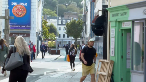 The image shows St Helier high street on a dry day. Members of the public are walking along the street to visit the shops.