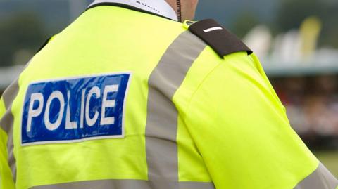 The back of a policeman wearing a large yellow high-viz jacket with grey patches on it. There is a label on it with 'police' written in white capital letters on a blue background