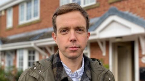 A man with short, dark brown hair wearing a green coat over a striped shirt stands in front of houses.