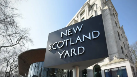 A stock image of the famous triangular New Scotland Yard sign outside the Metropolitan Police headquarters on a sunny day with a blue sky in the background.