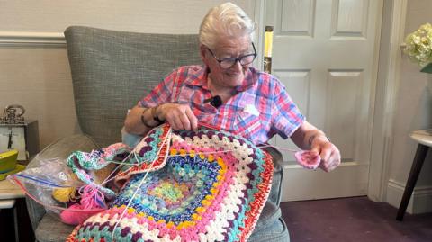 A woman with short white hair and glasses wearing a light ink and blue check short sleeved shirt is sat on a large grey armchair. On her lap is a crocheted blanket, beside her are a few balls of wool and she has a small square of the crocheting in her hands.