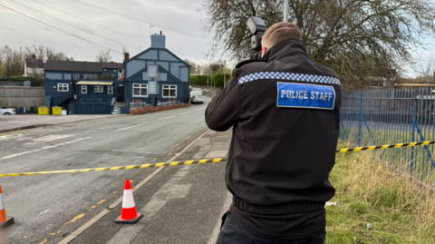 A person in a black jacket with the words police staff on the back is taking pictures from the side of a road. There are cones and yellow and black tape across the road.