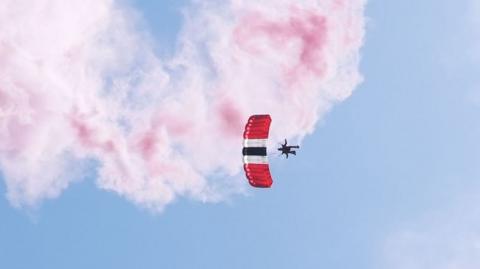 Image shows a parachutist in the sky with a red, black and white parachute. There is red smoke behind him. 
