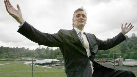 A man wearing a black pinstriped suit, yellow tie and blue and shite striped shirt is sitting with his arms outstretched. Behind him, down a hill, a stage is in the beginning stages of construction in front of a large grassy area, with trees behind it. It is an overcast day. 