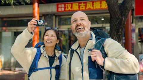 A man with a beard stands next to a woman with long, dark hair.  They are wearing matching beige, fluffy jumpers. They are looking up and the woman is taking a photo on a digital camera. There are Chinese characters on a sign above their head.