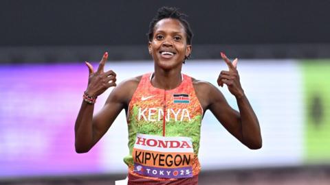 Faith Kipyegon, wearing an orange and green running vest, holds both index finders up towards her face as she smiles in celebration after winning a race