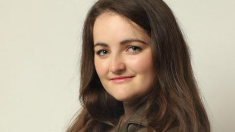 A woman with long brown hair and wearing a dark top, smiles as she looks to her left at the camera. 