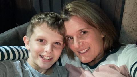 A mum with blonde hair and her tween son, with a similar hair colour, smile in a selfie together. The mum has her arm around her son. She is wearing a white, pink and black striped jumper, and her son is wearing a grey T-shirt.