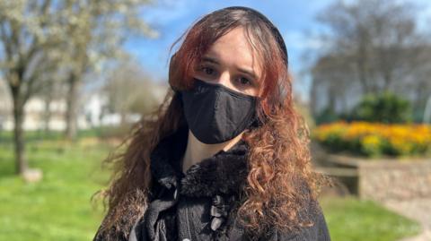 Stella Black with long curly red hair, wearing a black mask and headphones whilst stood outside in a park on a sunny day.