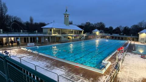 An outdoor swimming pool at twilight, with clear blue water and snow sprinkled around its perimeter