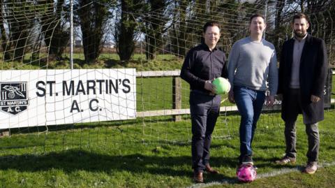 Three men stand in a football goal on a grass pitch - one holding a ball and one standing on one - with a St Martins AC banner behind them.