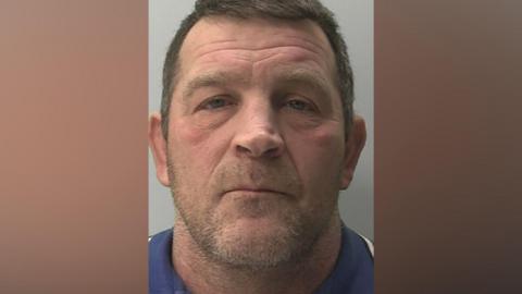 A mugshot of Paul Quinn, who is wearing a blue top, has light stubble, and is staring vacantly forward. 