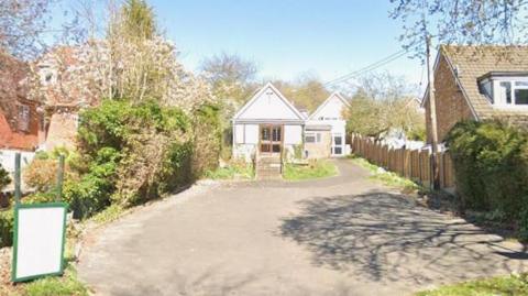 A close-up of the empty church surrounded by houses. The sky is blue with a tree casting a shadow on the driveway in the foreground