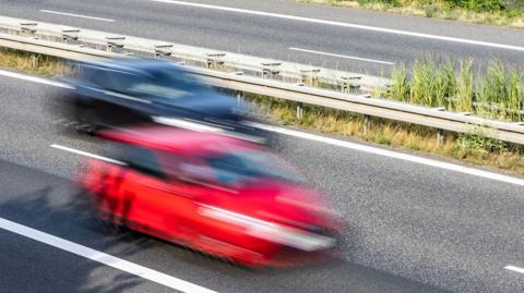 A stock image showing a blue and a red car speeding past the camera in a blur. They are driving along a dual carriageway with a metal barrier separating the lanes. 