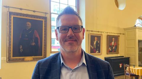 John Campion pictured at Pershore Civic Centre. He is wearing a navy blue suit with a light blue shirt. Behind his is a large window with portrait painting along the wall.