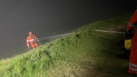 A man in an orange suit and a red helmet descends a grassy hillside in darkness