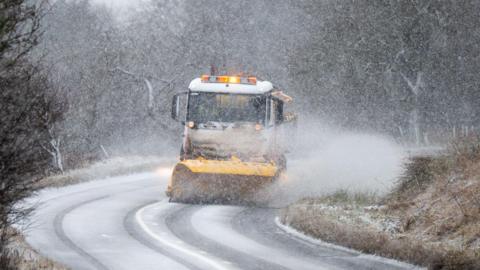 A snow plough travels round a bend on a single carriageway in the countryside, with snow swirling in the air and a thin layer lying on the ground.