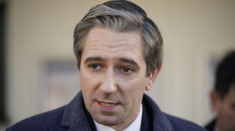 Simon Harris is wearing a navy coat. He is mid speech. He has grey white hair and brown eyes. His hair is curtain esque to the left. 