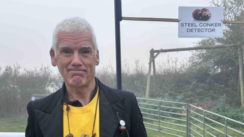 A man in a yellow t-shirt and black coat stands in front of a metal gate looking at the camera.