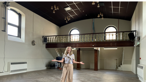 A woman stands in the centre of an historic chapel. She is standing on parquet flooring and behind her are two large windows and a raised area - which is accessed by two sets of stairs. She has her arms open side in greeting. 