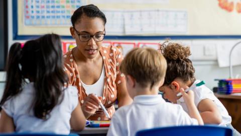 Primary school students sitting in a classroom being taught by a teacher. The pupils are learning on mini whiteboards.