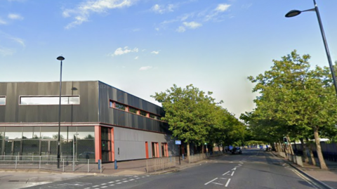 A large two-storey grey and red building is next to a road lined with trees.