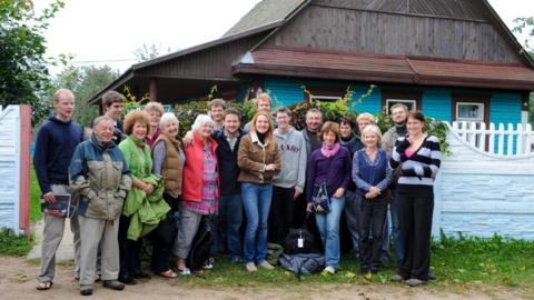 A group of people wearing jackets, jumpers and scarves stands outdoors in front of a wooden building and a pale blue fence in a rural setting, photographed in 2010.