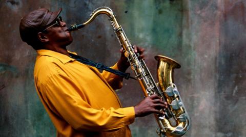 A musician leaning back as he plays on a saxophone. He is wearing a brown leather cap and a mustard coloured shirt. Behind him is a concrete wall.