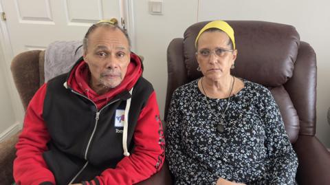 A man with a grey beard is pictured in a red and black hoodie while a woman sits beside him in a white living room wearing a black floral top and yellow headband.