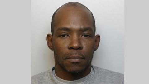 Police mugshot of David Malcolm. He is wearing a grey t-shirt and standing against a grey background.