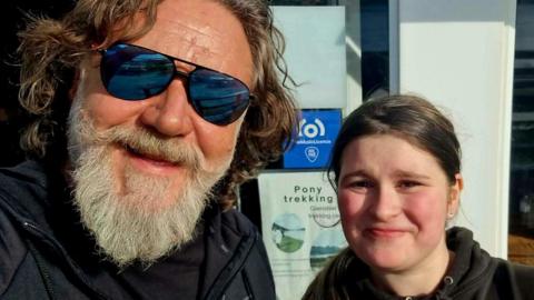 A man with a grey beard and shaggy brown hair. He's wearing mirrored sunglasses and is posing for a selfie with a young girl. She has dark hair and is wearing a black hoodie. They are both smiling. 