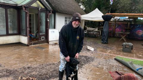 Andy Matthews and his dog Lokie. They are standing on flood-damaged outdoor ground. Debris is scattered in the garden. The exterior building of his home is behind him. Matthews is wearing a beanie hat and a hoodie. His dog is standing in front of him between his legs. Matthews is holing his ears. Benches are in the background of the garden.