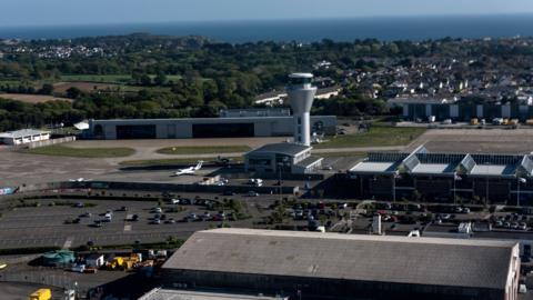 Aerial view of Jersey airport. A large car park, next to the airport, is half filled with vehicles. A wall separates the car park from the airport. Large buildings and an air traffic control tower overlook the car park. A plane is parked at the airport. Green trees and residential homes can be seen in the distance behind the site.