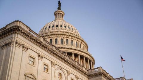 Exterior photo of the US Capitol, a beige building with a circular dome atop. An American flag is flying in the corner.