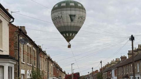 A hot air balloon over Bower Street in Bedford, showing roofs, brick terraced houses, telegraph cables, wires, lamp posts, in the sky, that is grey. There is a satellite dish on one building and a tree in the distance. 