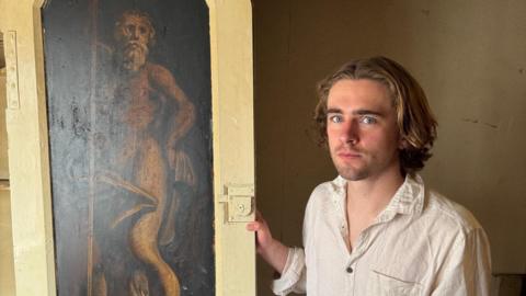 A man with brown wavy hair and a serious expression is wearing a off-white creased shirt. He is standing next to a sepia-coloured painting of an old man in a thick off-white cupboard door frame.
