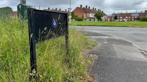 A black metal frame where the Dungeon Lane road sign would have been is surrounded by long grass. A main road runs alongside the lane with an terraced houses in the background.