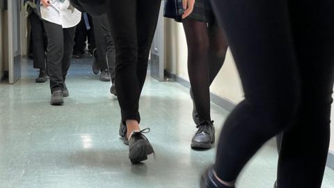 The photo shows the legs of secondary school children walking down a school corridor along a grey vinyl floor.