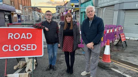 East Marsh councillors Nicola Aisthorpe, Steve Beasant and Loyd Emmerson, standing in front of railings and a road closed sign and cone at the end of a street with shops.