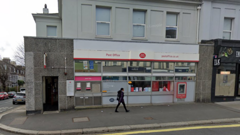 Photo of the Mutley Post Office, a grey building on a street corner, with a woman walking in front on the pavement